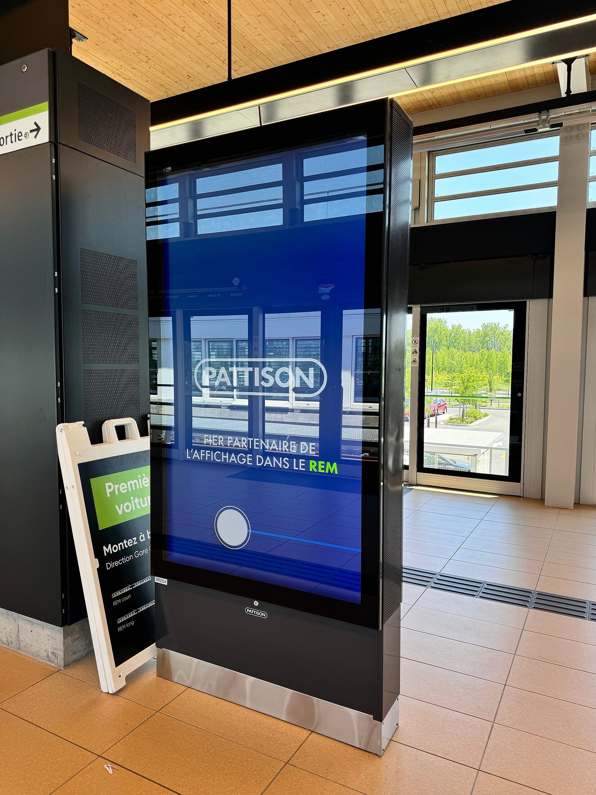 A tall portrait digital advertising screen standing in a railway station concourse